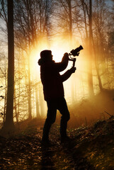 Silhouette of a filmmaker being creative in a misty forest, dramatically backlit with warm gold color and rays of light behind him - 648207275