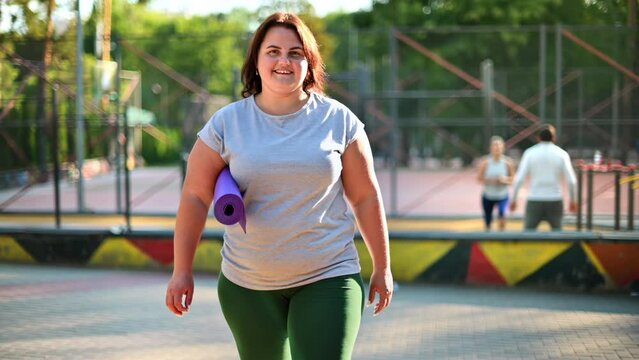 Woman with overweight in grey t-shirt and leggings walking on a sports field in a park while holding a yoga mat. Fitness lifestyle. Looking towards the camera