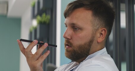 Male doctor sits in clinic cafe. Portrait of healthcare specialist talking by phone, recording voice message using social network or messaging app. Medical staff in hospital or medical center canteen.