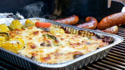Grilled vegetables and meat sausages on stainless BBQ grill with flames on dark background. Food and cuisine concept.