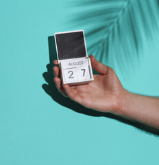 Man's hand holding wooden block calendar with date august 27 on blue background with palm leaf shadow