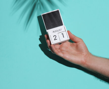 Man's hand holding wooden block calendar with date august 21 on blue background with palm leaf shadow
