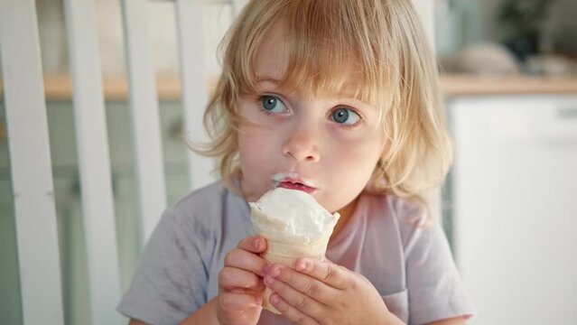 Baby girl enjoying ice cream. Pretty little toddler eating an ice-cream indoors, at home. Dining room background. Small child eats plombir and cream messy on her mouth. Cute kid with tasty sweet food.