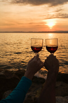 A couple holding wine glasses up at sunset over a lake; Bardolino veneto italy