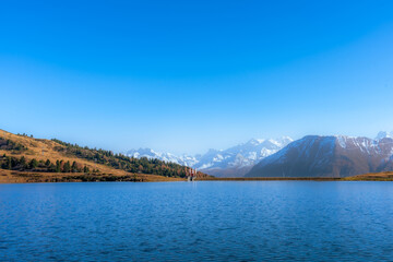 Scenic view of a lake, an autumn forest and mountains with snowy peaks.