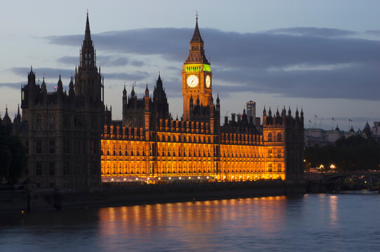 A Building And Clock Tower Along The Water's Edge Illuminated At Dusk; London, England
