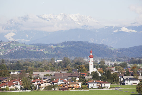 Alpine Village With Church Steeple And Rolling Hills With Snow Covered Mountains In The Background; Radfield, Austria