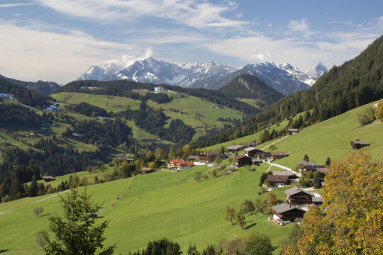 Alpine Village On Grassy Rolling Meadows With Snow Covered Mountain Range In The Distance With Blue Sky And Clouds; Alpbach, Austria