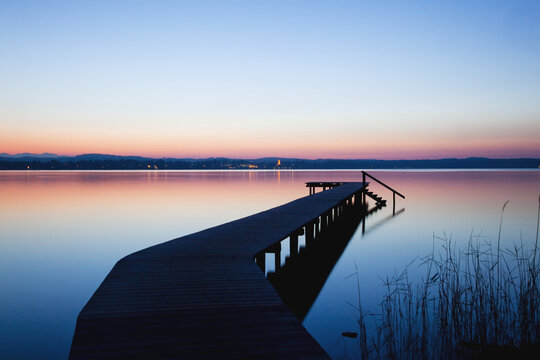 A Wooden Dock Leading Out To A Tranquil Lake At Sunset; Starnberger See, Bavaria, Germany