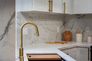 Modern kitchen details of gold faucet over sink with cutting board with full marble counter and backsplash.