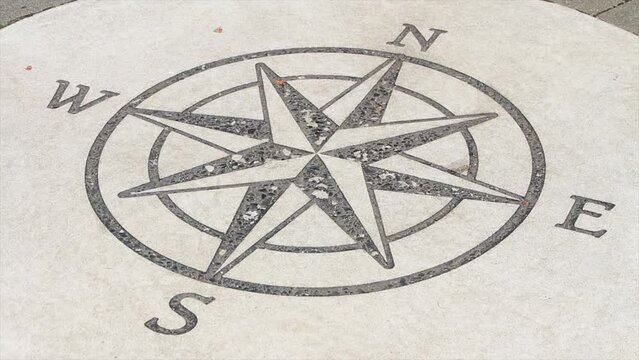 compass illustration print on circle cement pavement concrete slab in black with n e s w surrounded by gray brickwork floor, close up