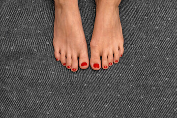When passion passes through the foot. Feet of a young girl with fiery red nail polish on gray and white uniform textured background. Neutral space and focus on beautiful well-groomed female legs. - obrazy, fototapety, plakaty