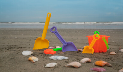 Beach toys, shovels and bucket on the sand surrounded by shells with the sea and blue sky in the background