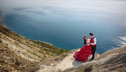 Wedding photo shoot of a beautiful bride and groom in red dresses on a mountain overlooking the...