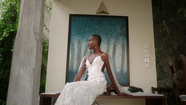 Portrait of gay black male in white wedding dress posing, smiling for fashion photo shoot. Elegant gender fluid bride sits, turns to camera against abstract blue picture in luxury retro villa.