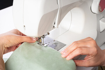 Tailor hands stitching green fabric on modern sewing machine at workplace in atelier. Women's hands sew pieces of fabric on sewing machine closeup. Handmade, hobby, repair, small business concept