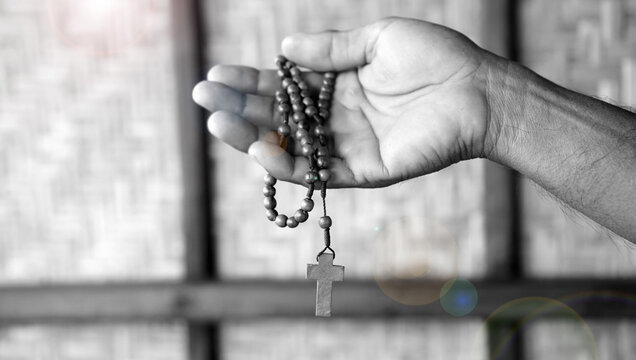 Person holding a wooden rosary in hand in black and white background with light. Praying rosary concept, catholic symbol of devotion to Mother Mary.