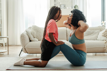 African American woman doing sports at home, little daughter helping, mother do abs in living room