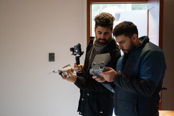 Two men using a drone and a compact camera inside a hotel room. Copy space available.