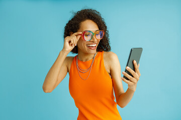 Smiling woman in eyeglasses looking on mobile phone while standing on blue studio background