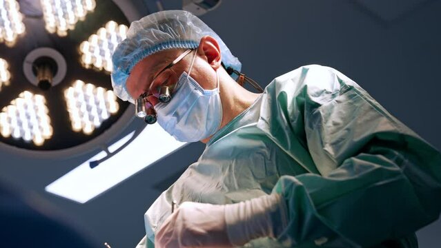 Focused adult male surgeon performs operation with metal tools. Portrait of a doctor in device glasses at surgery. Low angle view.