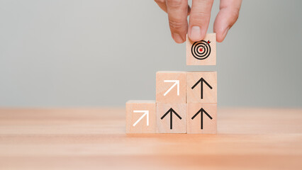Male hand put a wooden cube block with the goal icon on the top of stacking wooden cubes standing with arrow up icon. Business concept of goal achievement, financial growth, target and investment.