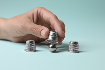 Woman showing metal ball under thimble on light blue background, closeup. Thimblerig game - obrazy, fototapety, plakaty