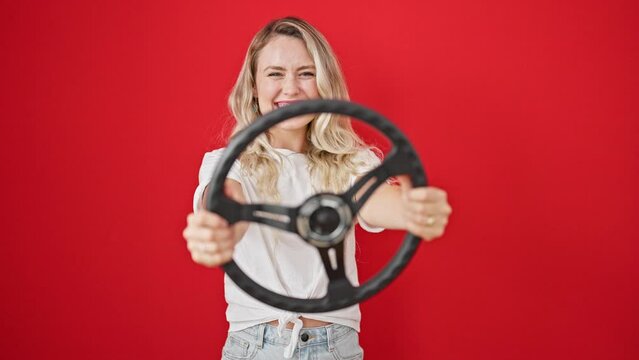 Young blonde woman smiling confident using steering wheel as a driver over isolated red background