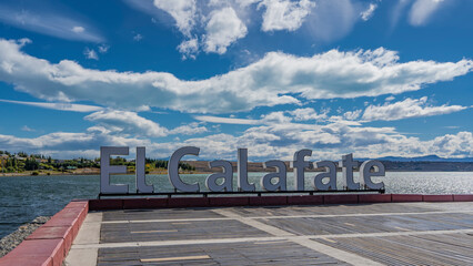 On the city embankment, the name of the city of El Calafate is set in large letters. Behind is the turquoise lake  Lago Argentino. Clouds in the blue sky. Argentina.