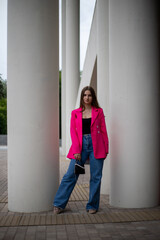 Fototapeta na wymiar a young business girl in a bright fuchsia jacket and jeans stands at full height on the street with a small handbag