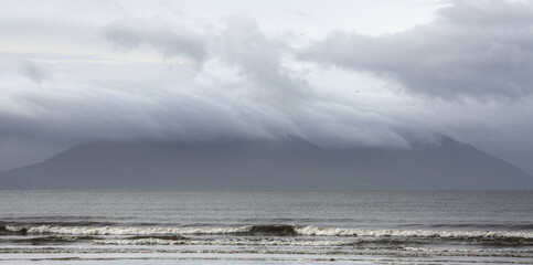 A mountain is obscured by morning storm clouds, which produce a blueish-gray scene with soaring birds, a dramatic sky, and crashing waves. Inch Beach, Dingle Peninsula, County Kerry, Ireland