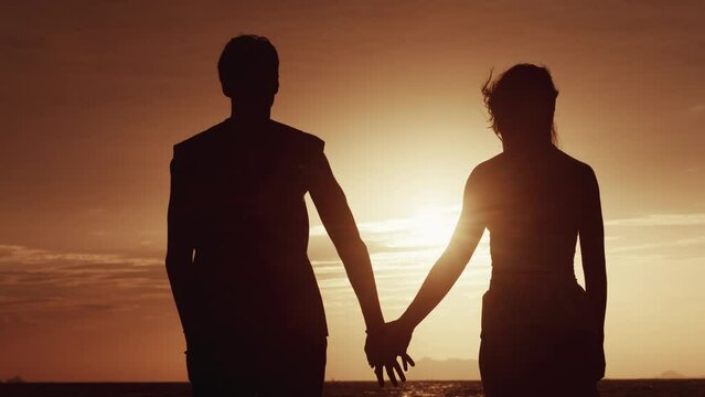 Back view, a man takes a woman's hand against the backdrop of an orange sunset. Silhouette of a loving couple at sunset at sea while traveling on honeymoon. The concept of a man's love for a woman