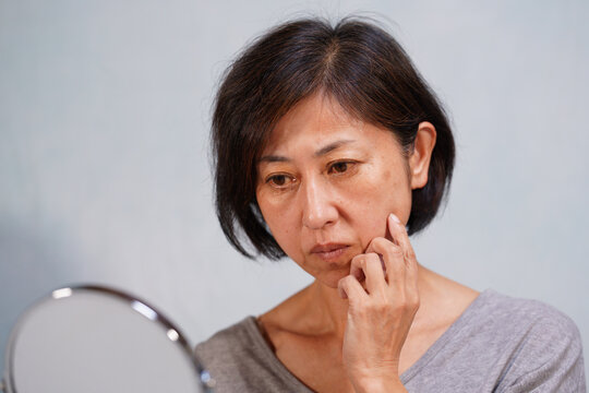 鏡を見ながら頬に手をやるシミ、シワのある老け顔の中年女性