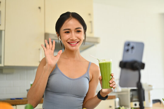 Confident female vlogger holding glass of fresh green detox vegetable juice and filming vlog about healthy food in kitchen