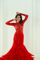 Brunette woman with makeup and long curly hairstyle wearing red evening gown in white interior. Vogue style model