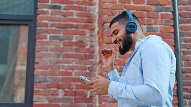 Side view of carefree man of indian ethnicity walking along brick building and listening to favourite music through modern wireless headphones. Positive person creating own track list on smartphone.
