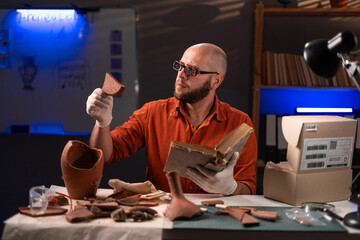 Archaeologist working at night in the office studying parts of an antique vase, archaeological...
