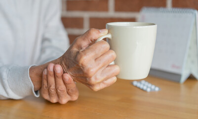 close up senior woman hold on hand to relief shaky symptom while drink water for Parkinson’s...
