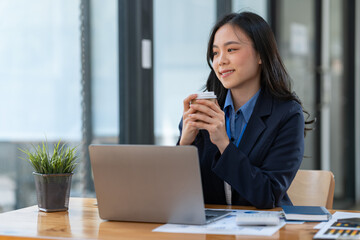 Attractive businesswoman holding cup of hot drink and looking away with smile while sitting in office.