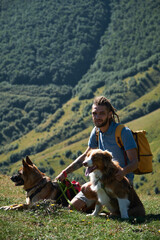 A tourist trip to Georgia. Caucasian young guy with dreadlocks sits on top of a mountain in summer with two dogs. The male owner travels with Australian and German Shepherds.
