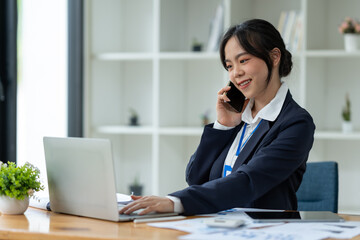 Asian businesswoman using mobile phone to communicate, chat, negotiate, search internet, news, email while working online on laptop computer at office.