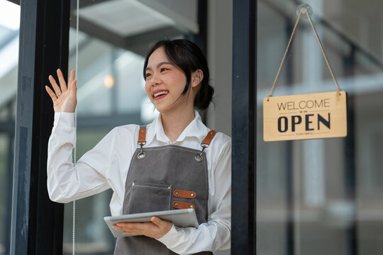 Young freelance businesswoman Asian young female barista in apron holding sign preparing to open for sale and standing in front of the door of the cafe with open sign business owner startup concept