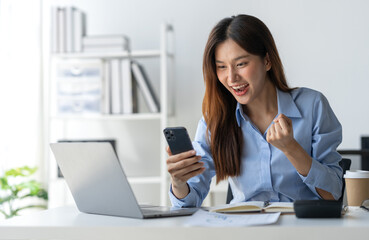 Fototapeta na wymiar Asian businesswoman raising her hand with a happy expression and looking at her phone The businesswoman was delighted to receive an email informing her of her annual bonus. concept of success