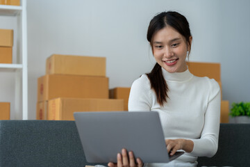 Asian businesswoman taking orders and checking online from laptop and mobile phone to prepare merchandise and deliver to customers in home office. business concept sme e-commerce freelance.