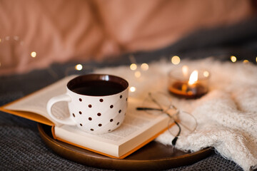 Cup of tea stay on open paper book with scented candle on knit clothes in bed close up. Winter...