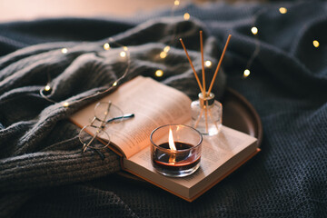 Scented burning candle in glass jar with glasses on open paper book and diffuser in bed over glow...