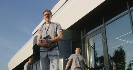 Fototapeta premium Mature doctor with tablet in hands stands with patient at clinic entrance, discuss treatment. Healthcare specialist says goodbye to client after appointment, then looks at camera. Medic works outside.