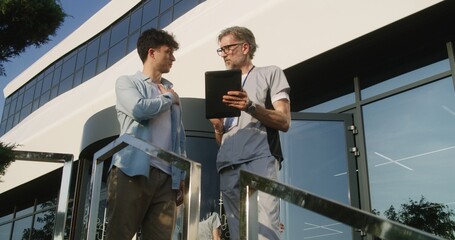 Mature doctor with digital tablet in hands stands with adult patient at clinic entrance and discuss his treatment. Health care specialist consults client or makes diagnosis. Physician works outside.