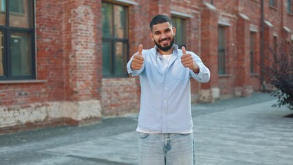 Portrait of handsome man expressing contentment and happy emotions while resting on fresh air and raising thumbs up. Cheerful male dressed in jeans and sky blue shirt showing gestures of approval.