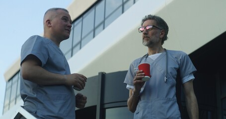 Two doctors in uniform stand near medical center entrance in the evening. Professional medics drink coffee during break and talk. Medical staff of hospital or modern clinic. Lifestyle and healthcare.